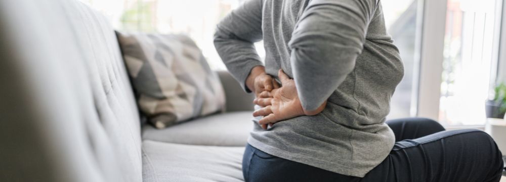 ALT: Woman sitting on couch holding back with both arms due to pain 